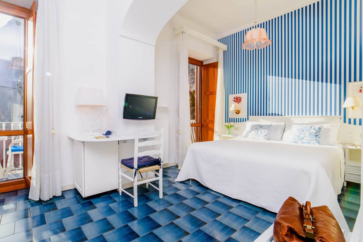 A hotel room with a bed, a desk with a TV, a chair, and a suitcase.