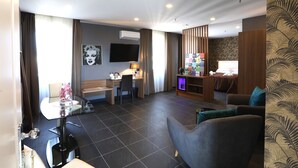A hotel room with a bed, a seating area with a sofa and armchairs, a desk with a chair, a TV, and a small entertainment unit.