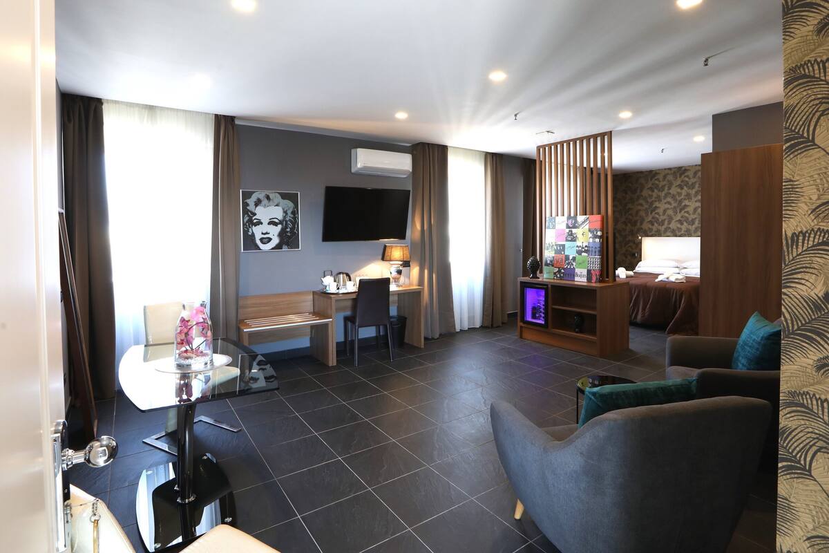 A hotel room with a bed, a seating area with a sofa and armchairs, a desk with a chair, a TV, and a small entertainment unit.