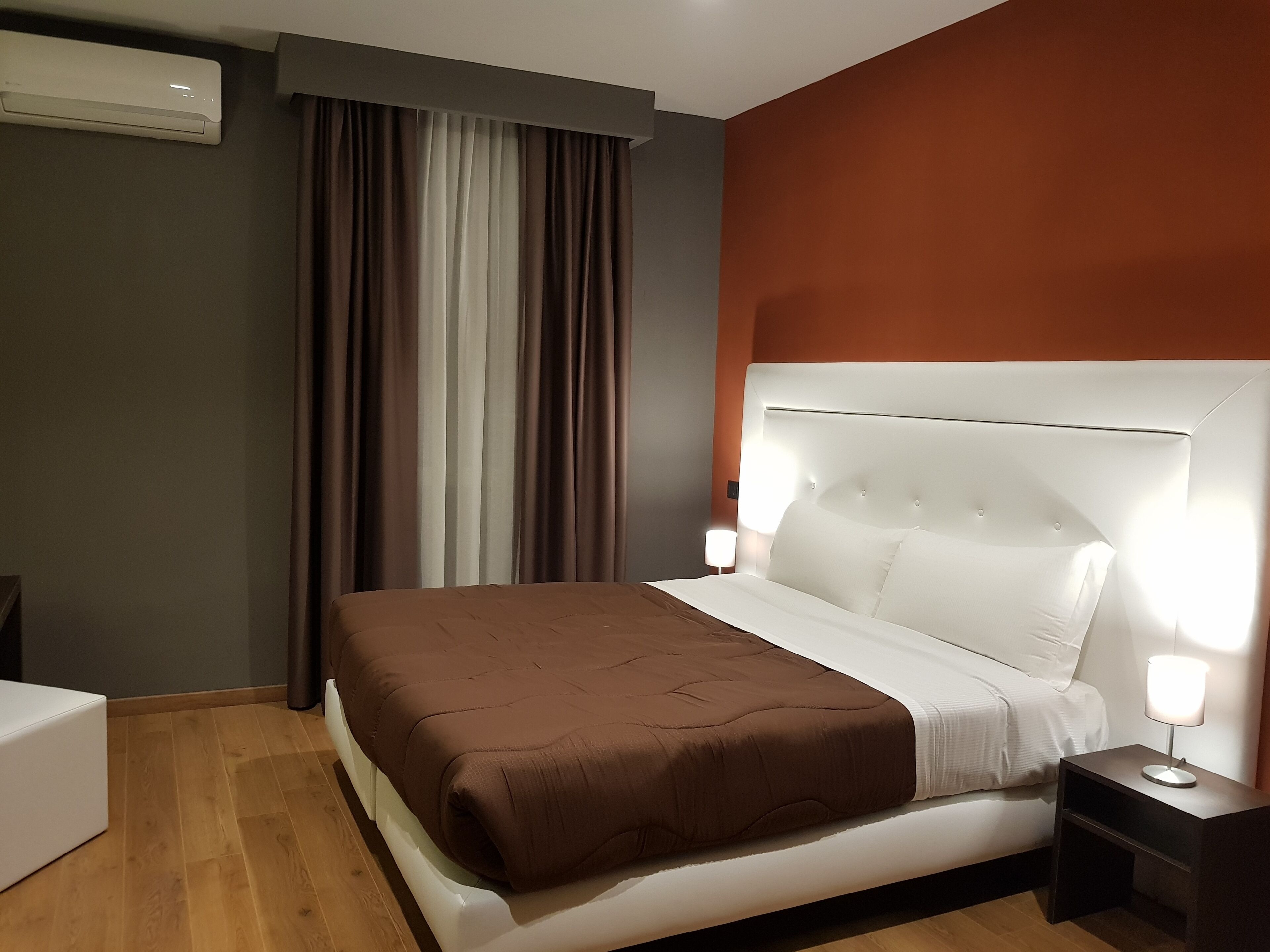 A hotel room with a bed, a bedside table with a lamp, a window with curtains, and an air conditioning unit on the wall.