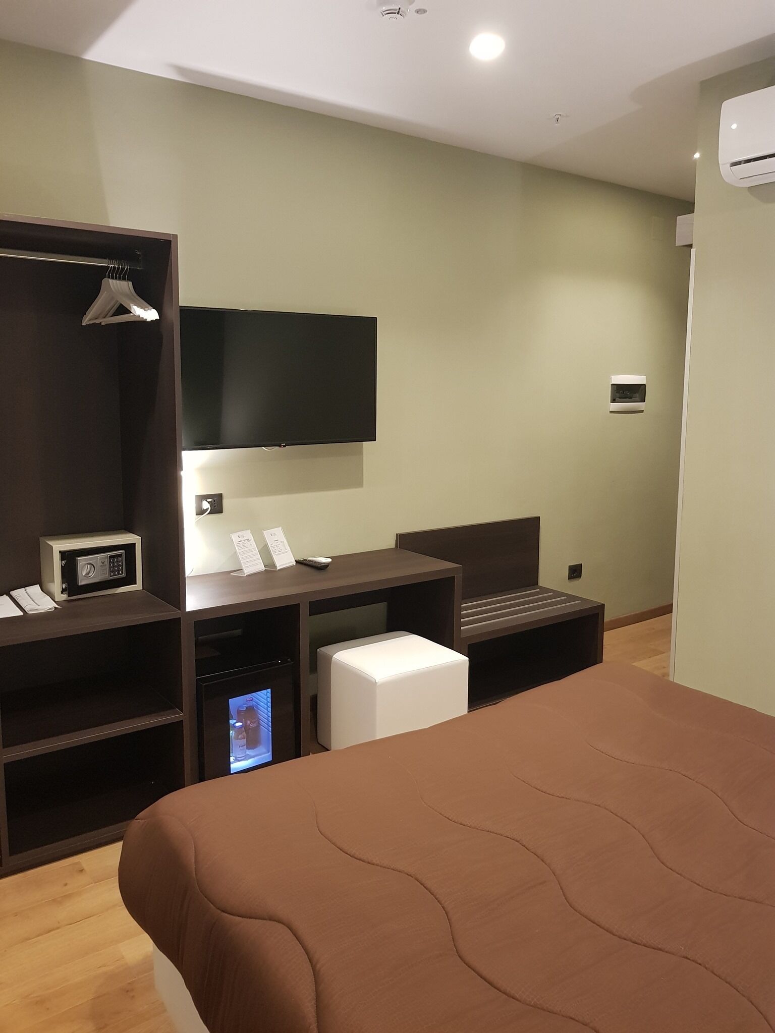 Una habitación de hotel moderna con una cama grande, un televisor de pantalla plana montado en la pared, un escritorio con computadora y una caja de seguridad pequeña, y un mini bar.