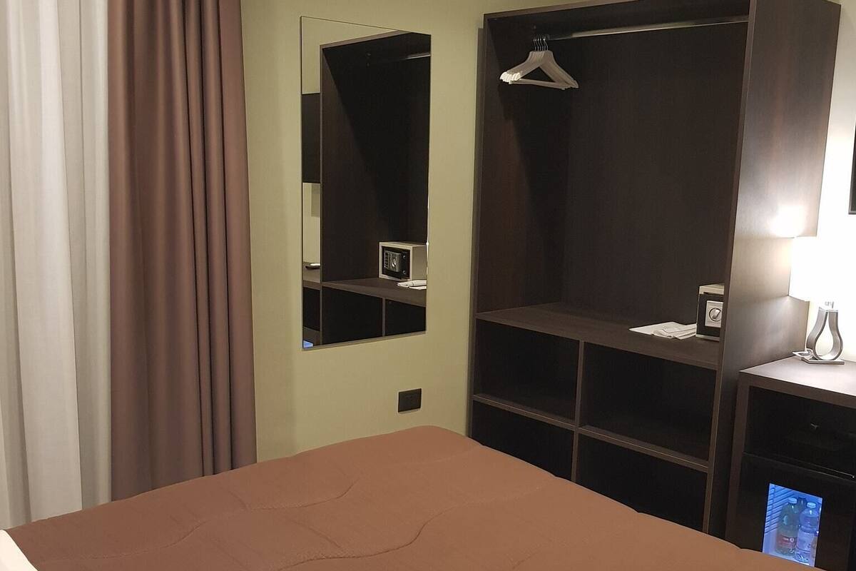 A hotel room with a bed, a wardrobe, a mirror, and a small table with a lamp.
