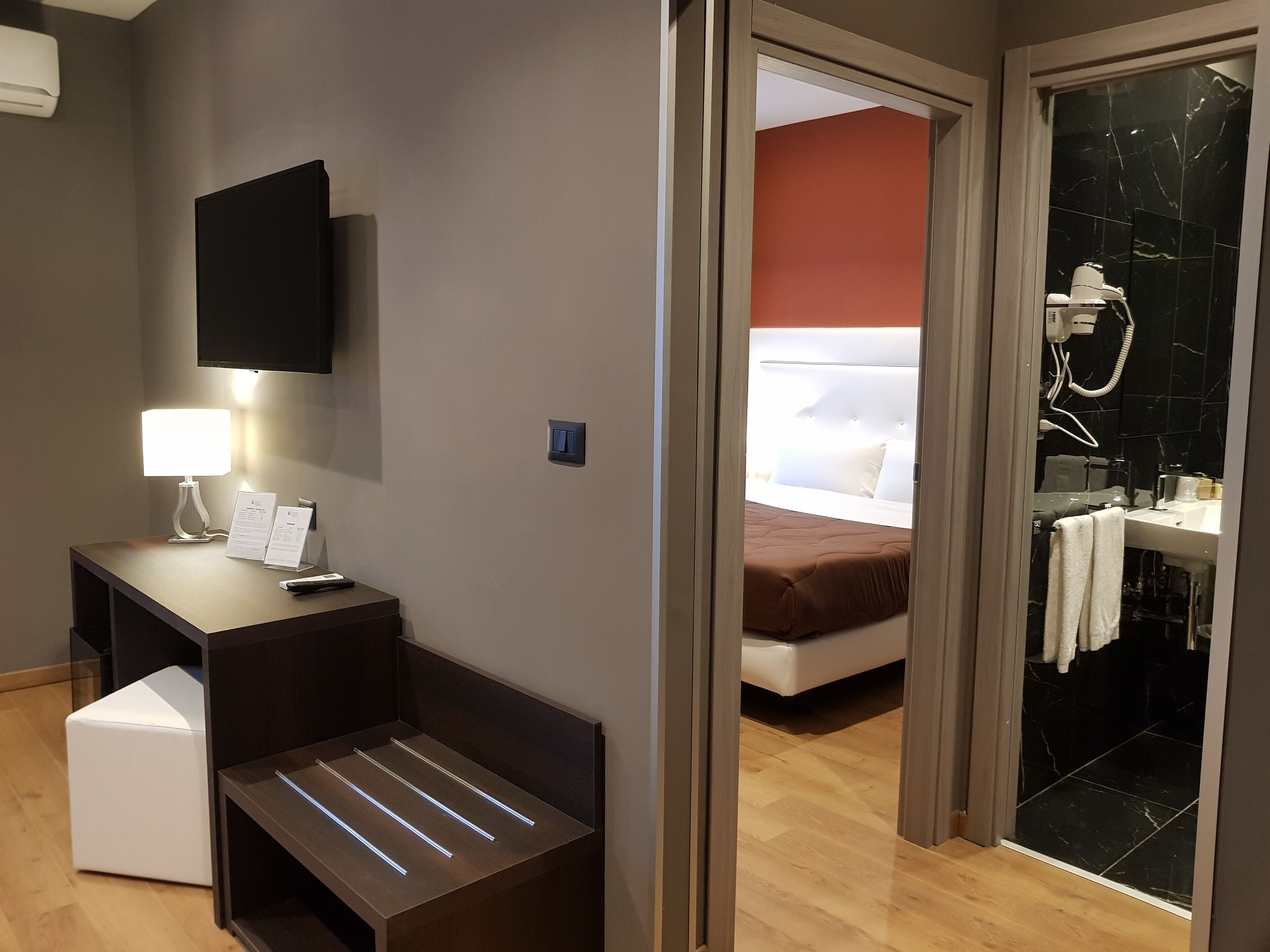 A hotel room with a bed, a bedside table with a lamp, a flat-screen TV, and a shower area with a glass door.