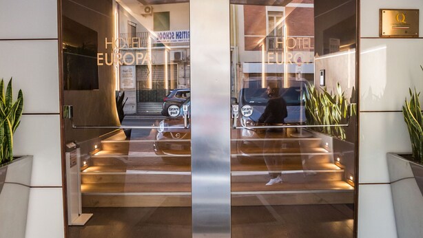 The entrance of HOTEL EUROPA with a modern design, featuring a glass door, a central pillar, and a logo above the entrance.