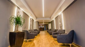 A hotel lobby with a wooden floor, grey armchairs, a large potted plant, and a reception desk in the background.