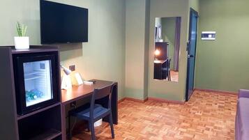 A compact room with a wooden floor, a desk with a chair, a television mounted on the wall, and a refrigerator with a potted plant on top.