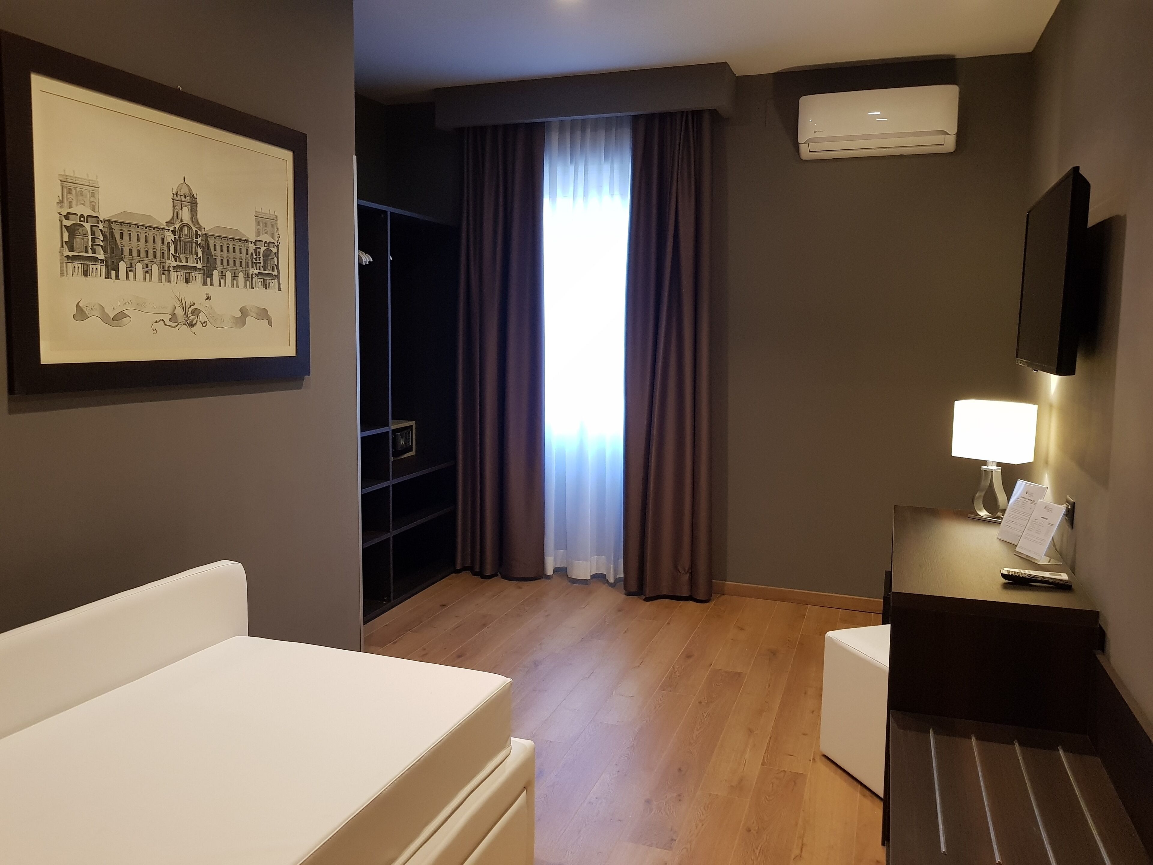 A hotel room with a bed, a desk with a lamp, a TV, a framed picture on the wall, and a window with curtains.
