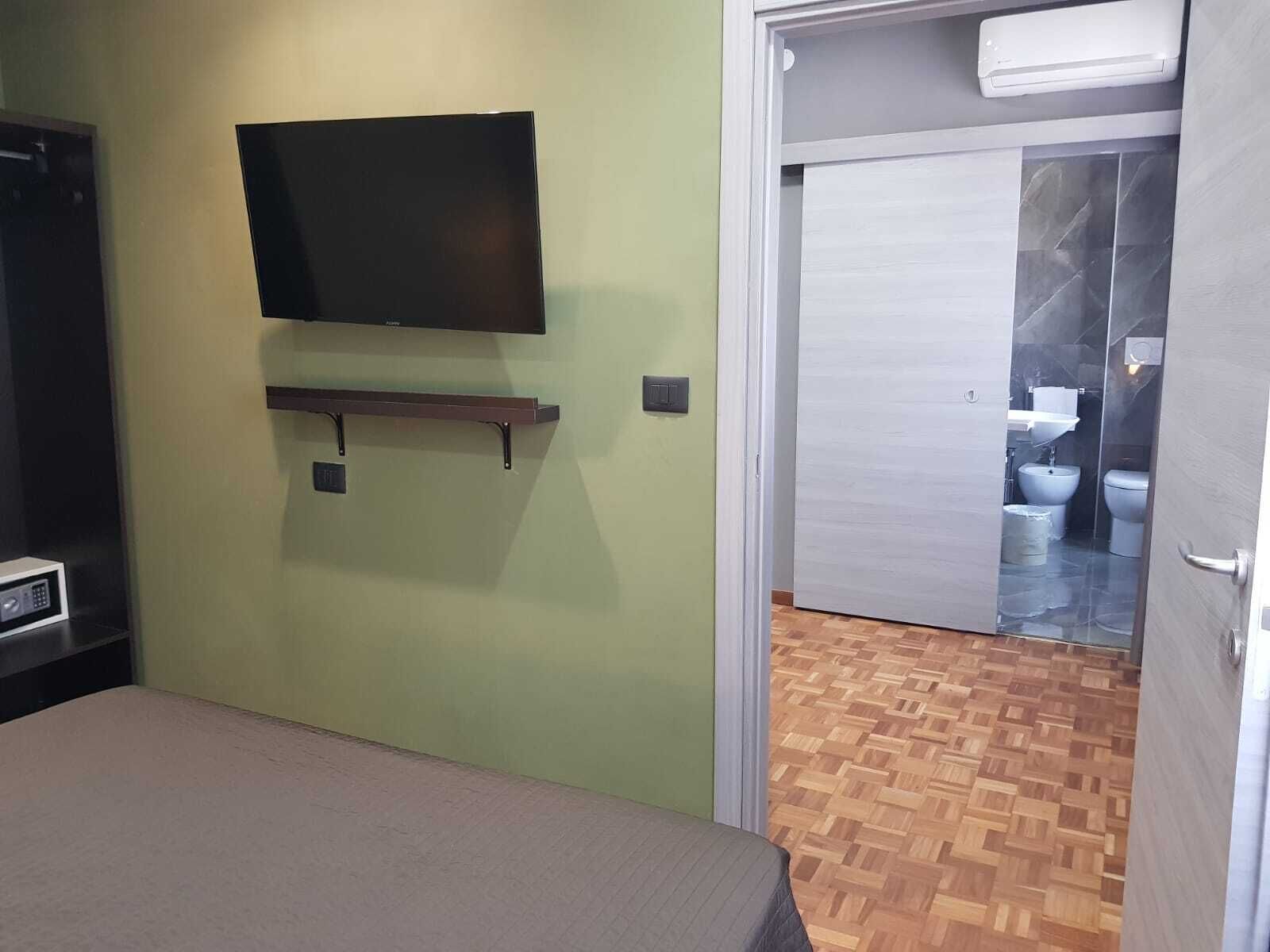 Une chambre comprenant un lit, un téléviseur fixé au mur, une petite étagère et une porte donnant sur une salle de bain équipée de toilettes et d’un bidet.