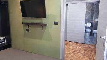 Une chambre comprenant un lit, un téléviseur fixé au mur, une petite étagère et une porte donnant sur une salle de bain équipée de toilettes et d’un bidet.