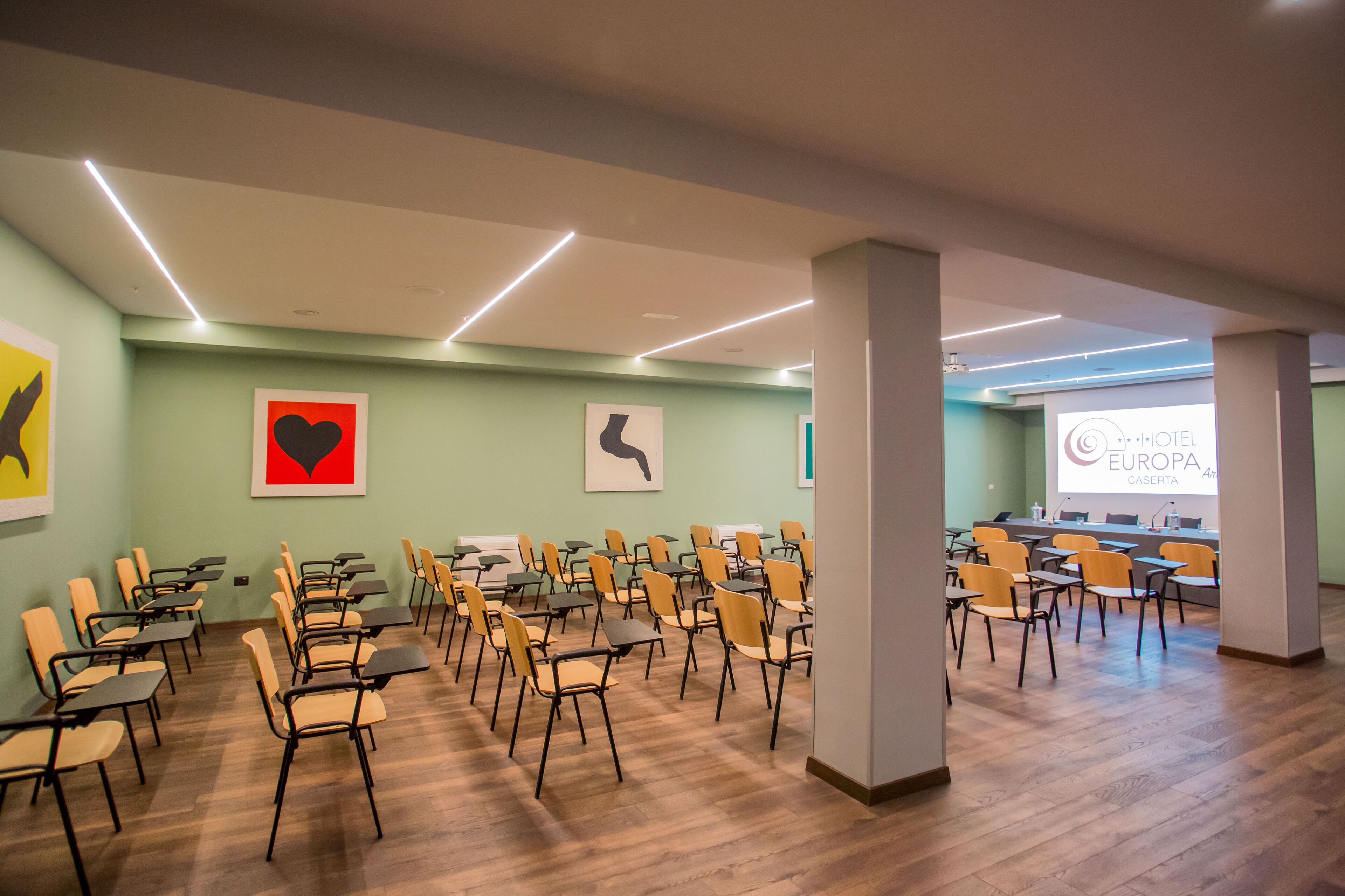 A conference room with rows of chairs and tables, a projector screen displaying 'EUROPA CASERTA', and artwork on the walls.