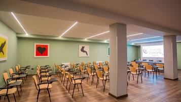 Une salle de conférence avec des rangées de chaises et de tables, un écran de projection affichant le texte « EUROPA CASERTA », et des œuvres d’art accrochées aux murs.