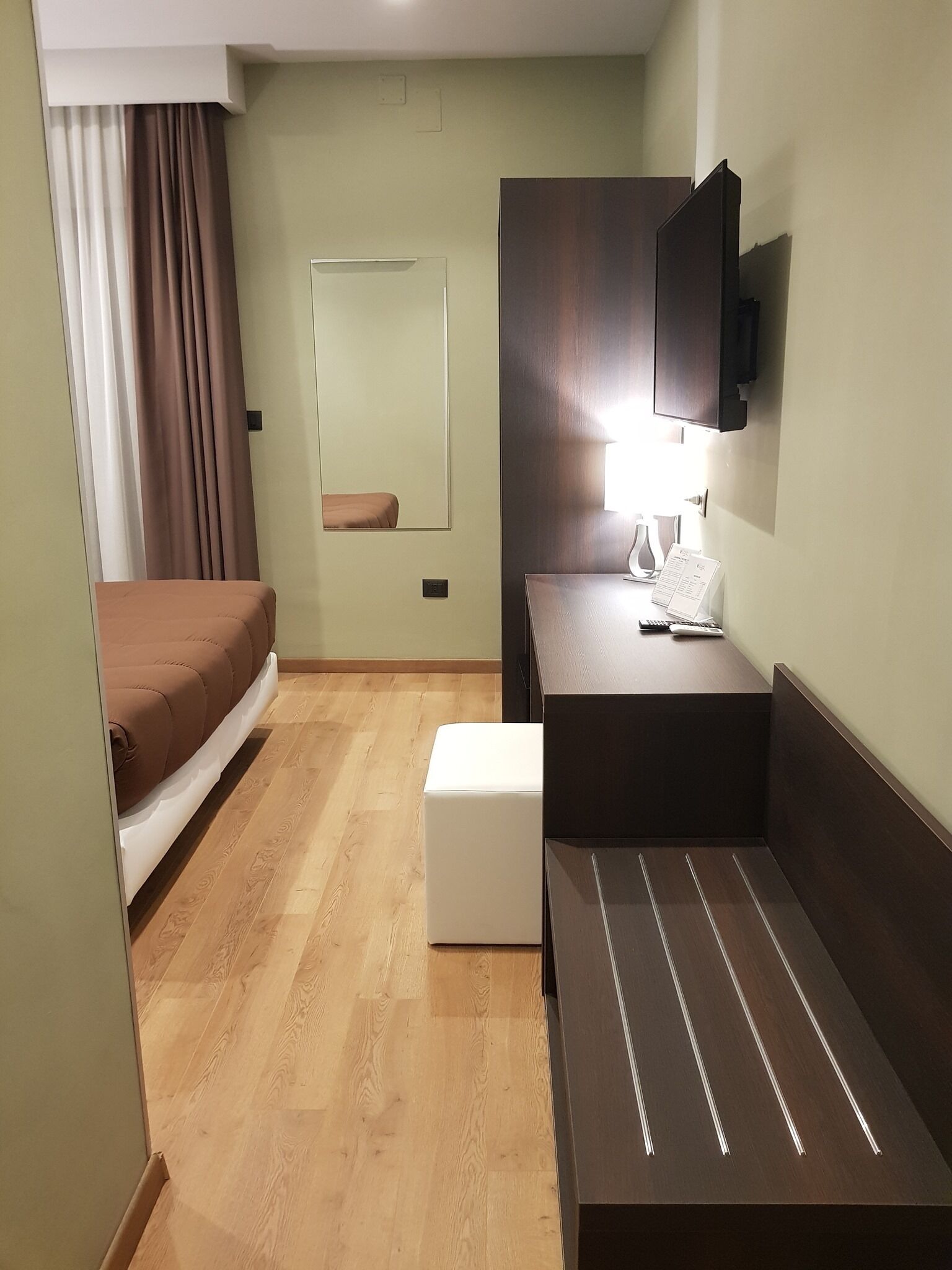 A hotel room with a bed, a bedside table with a lamp, a mirror, and a small white ottoman.