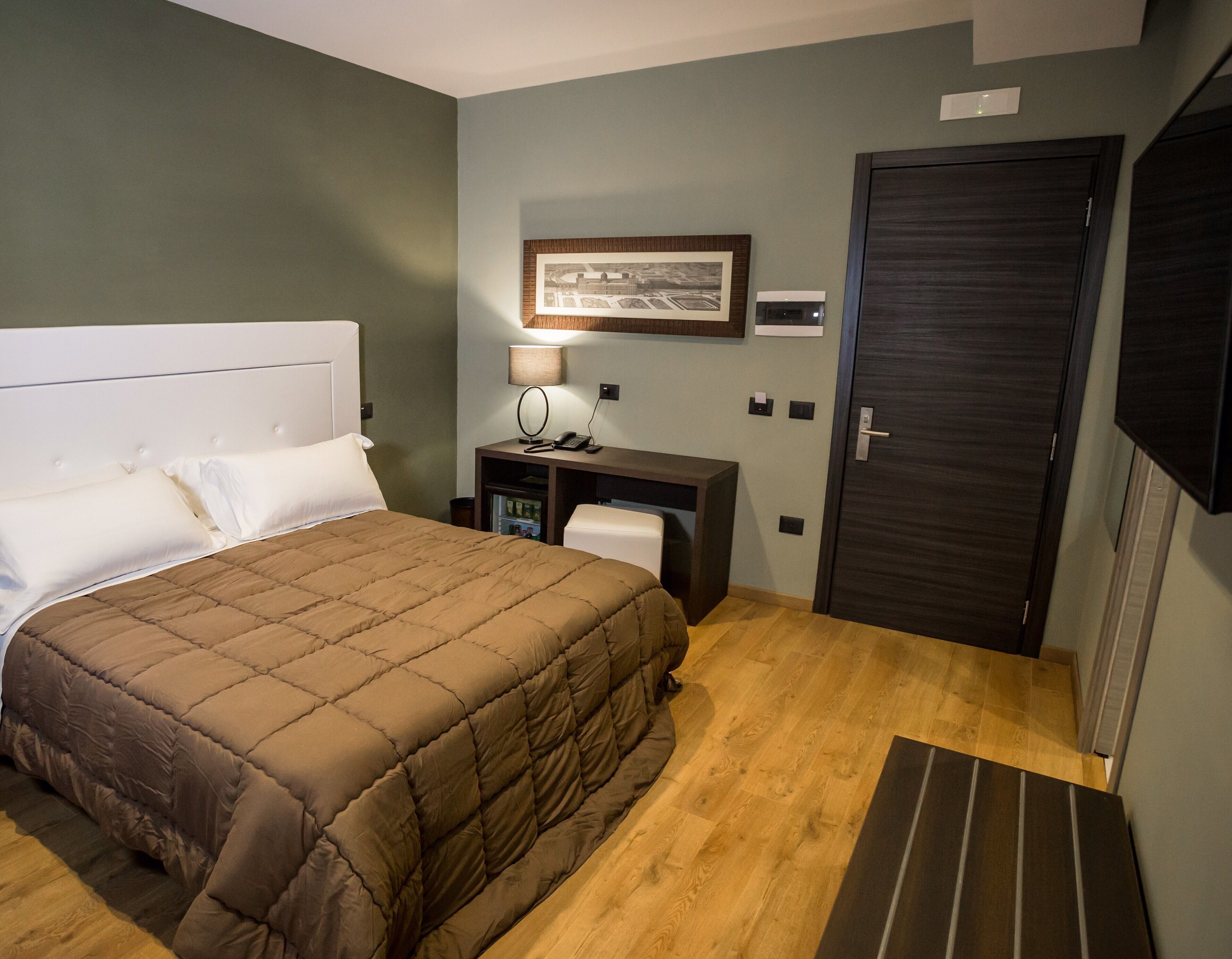 A hotel room with a large bed, a desk with a lamp, a framed picture on the wall, and a door.