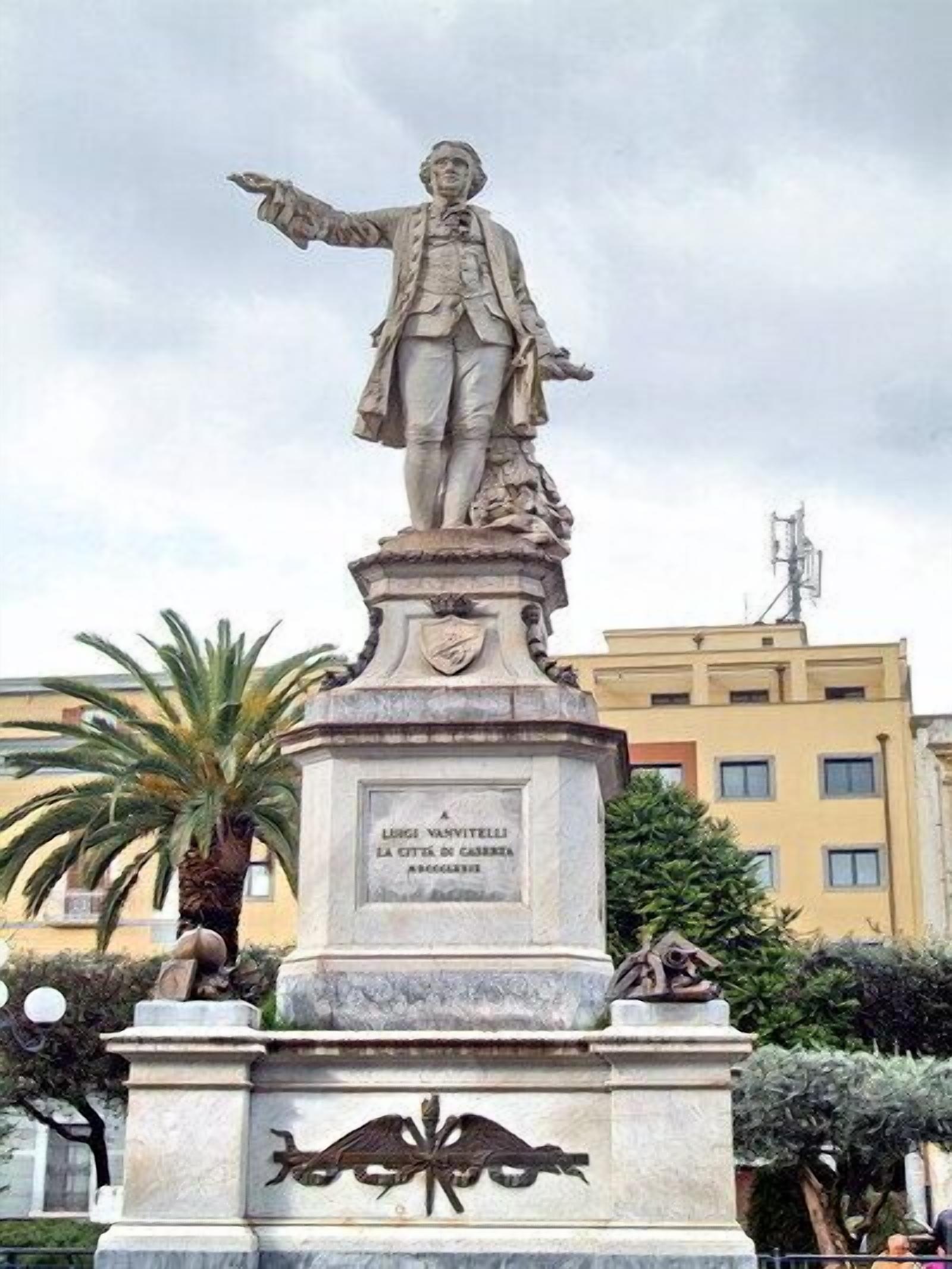 Una estatua de una figura histórica con una inscripción en su pedestal, ubicada en una plaza pública con edificios y árboles al fondo.