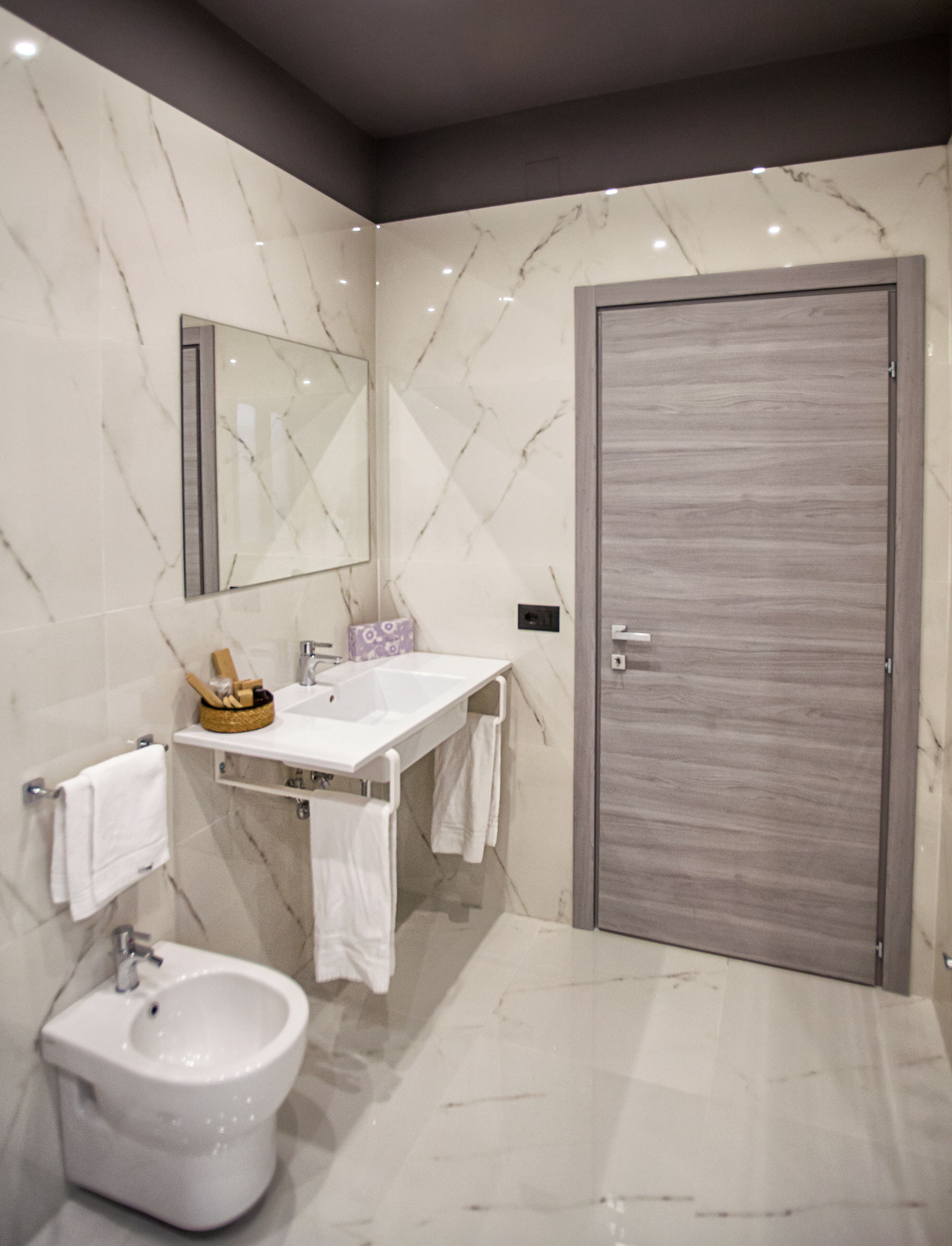 Un baño moderno con pared de mármol, un lavabo blanco, un bidé, un espejo grande y una puerta de madera.