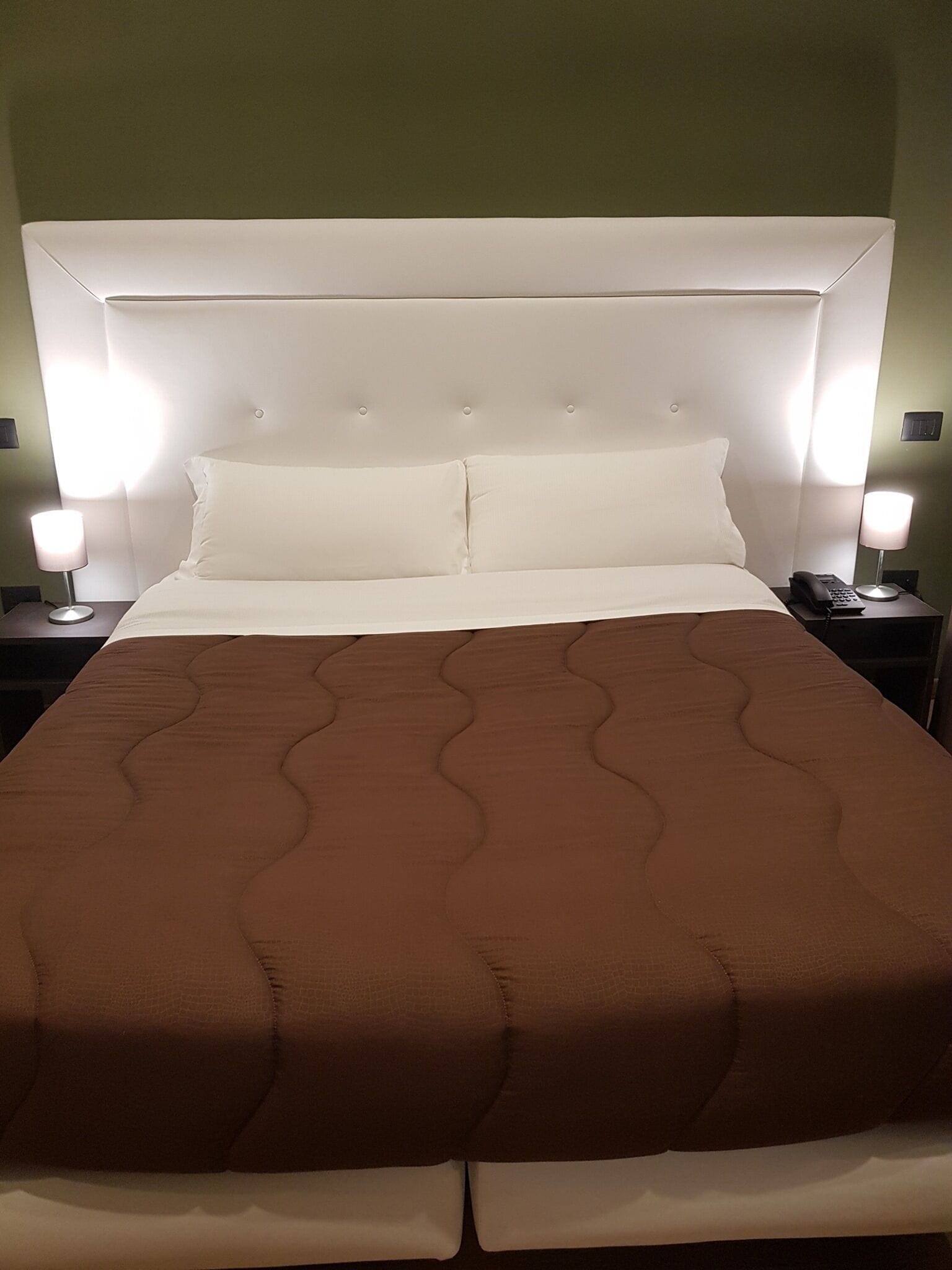 Una cama bien tendida con colcha marrón, almohadas blancas y un cabecero con una lámpara. Hay dos mesitas de noche, una con lámpara y teléfono.