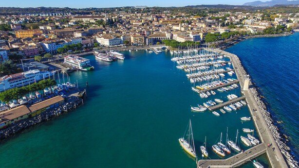 An aerial view of a marina with numerous boats docked, surrounded by a coastal cityscape with buildings and hills in the background. - Hotel City (Desenzano del Garda)