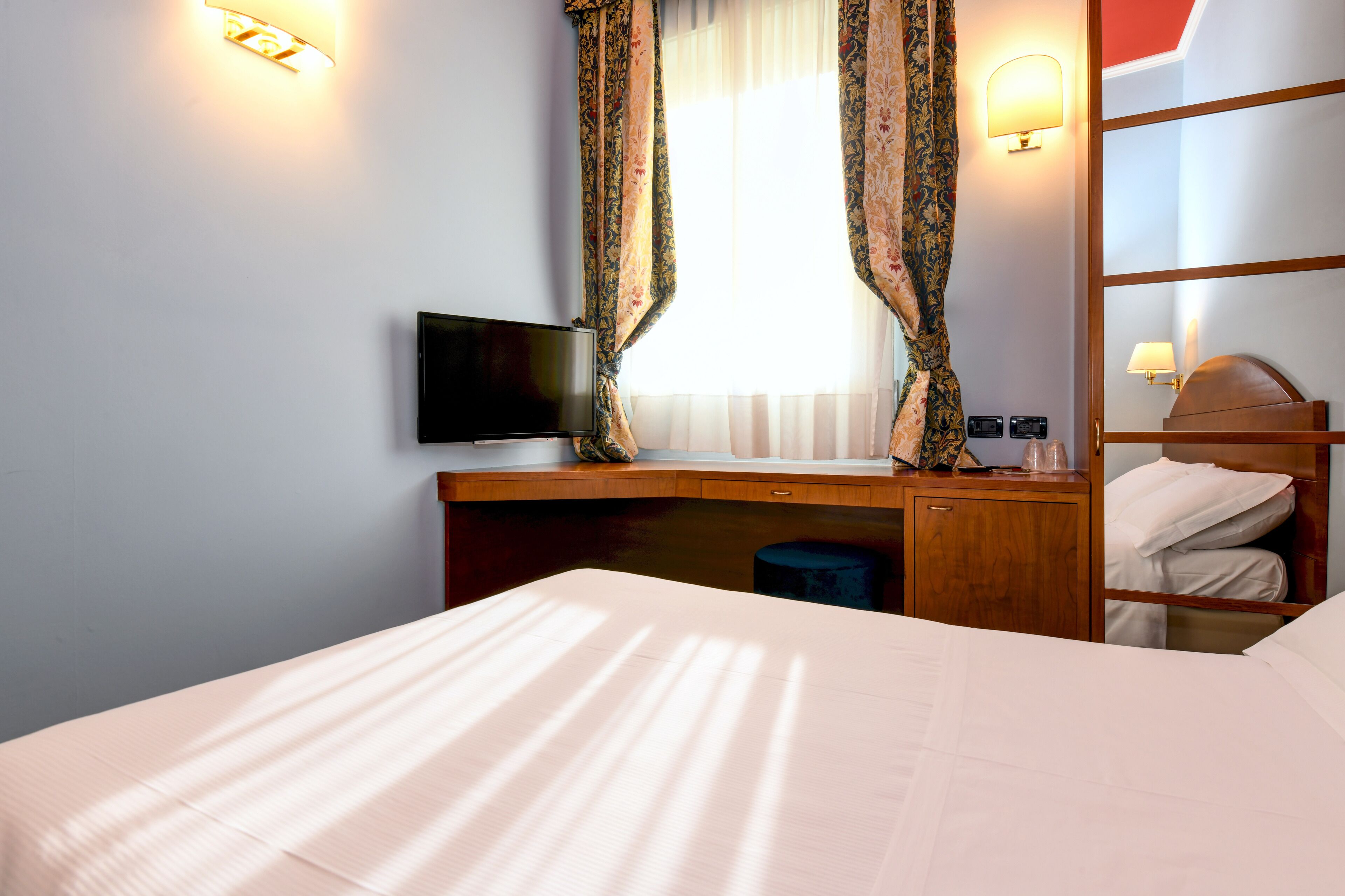 Una habitación de hotel con una cama, un escritorio con televisor, una mesita de noche, un espejo y cortinas.