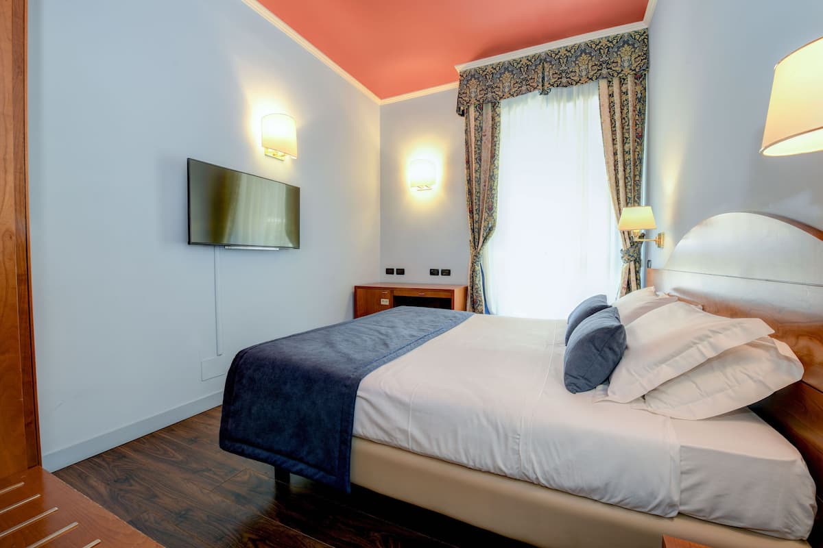 Een hotelkamer met een bed, een aan de muur bevestigde televisie, een nachtkastje en een raam met gordijnen.
