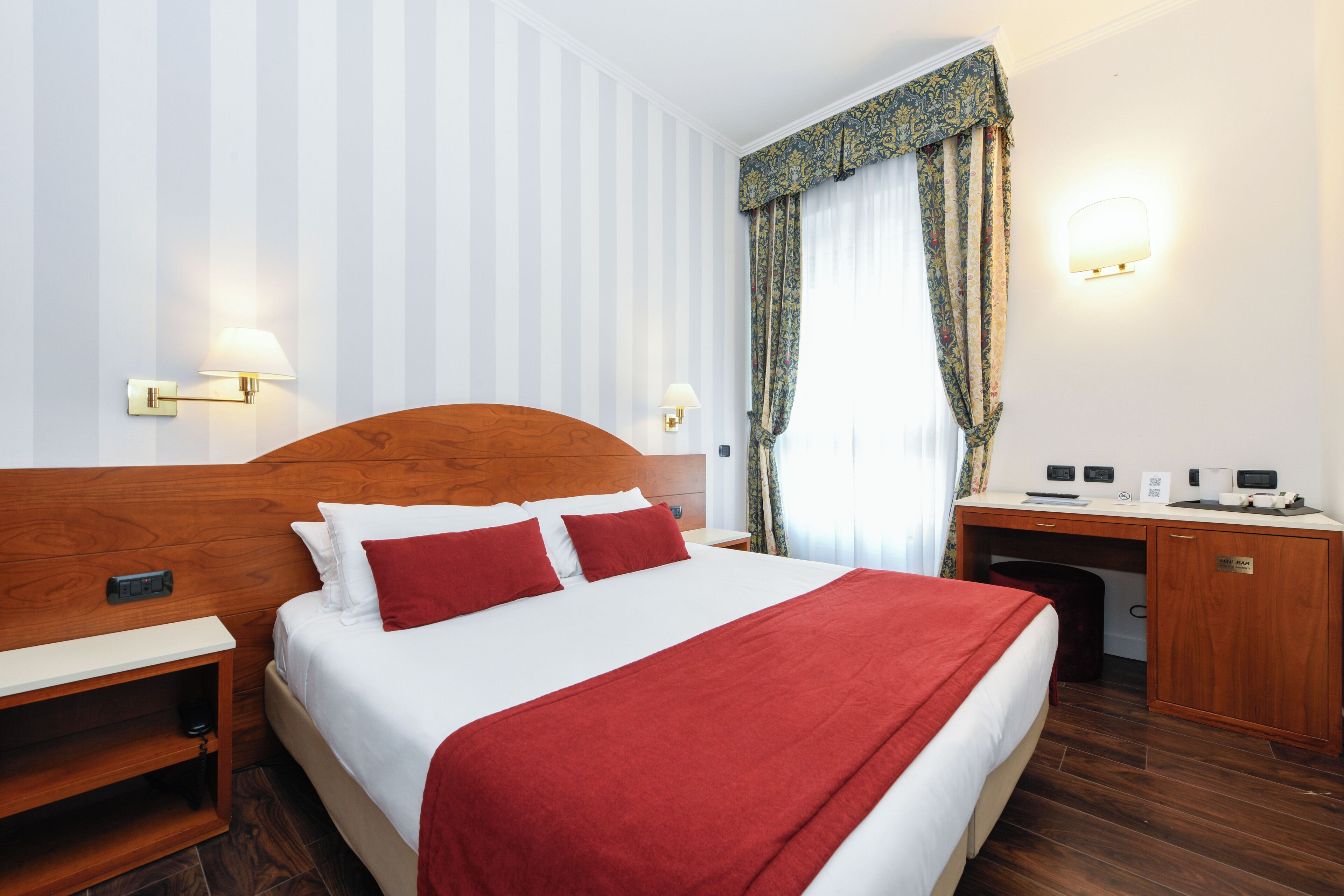 Ein Hotelzimmer mit einem großen Bett, einem hölzernen Kopfteil, einem Schreibtisch mit Computer und einem Nachttisch.