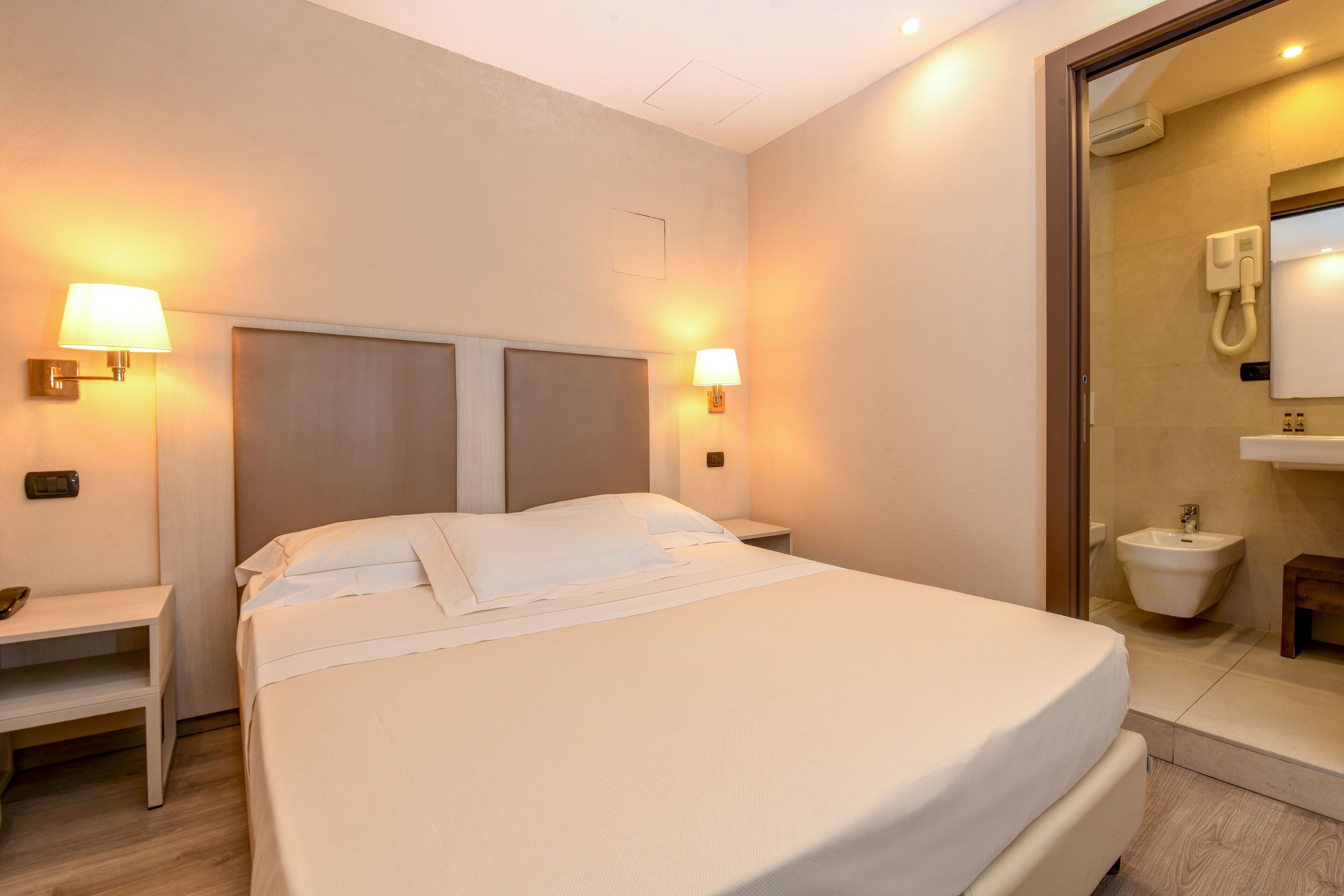 A hotel room with a large bed, bedside lamps, a nightstand, a mirror, and a bathroom with a sink and toilet.