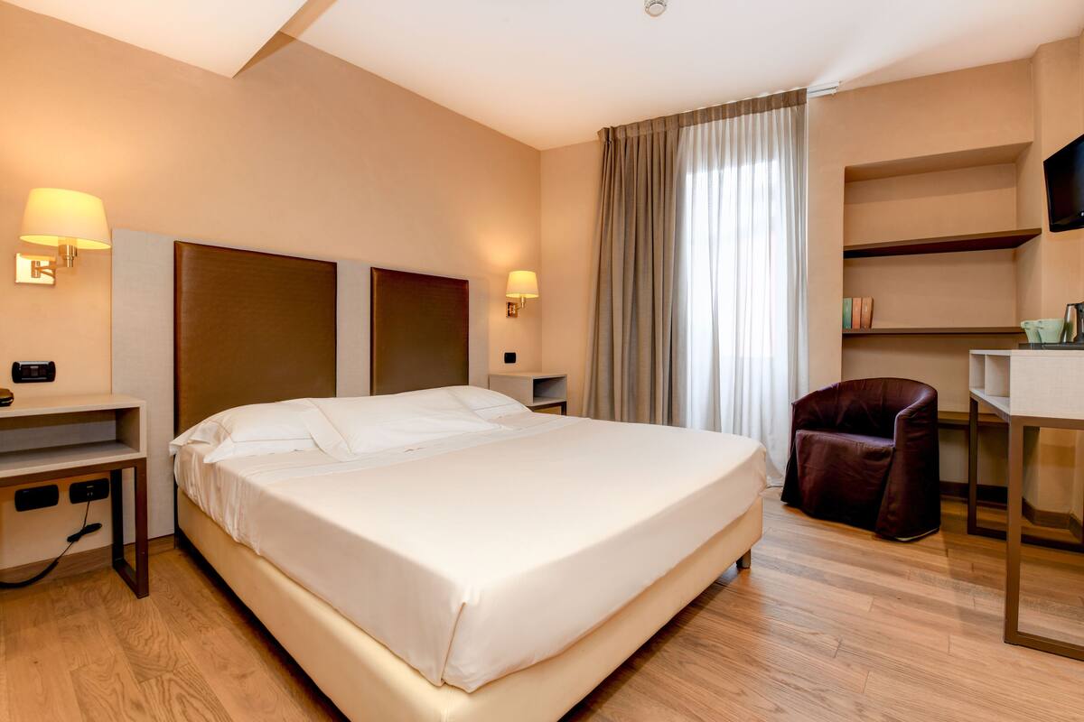 A hotel room with a large bed, bedside tables, a desk, a chair, and a TV mounted on the wall.