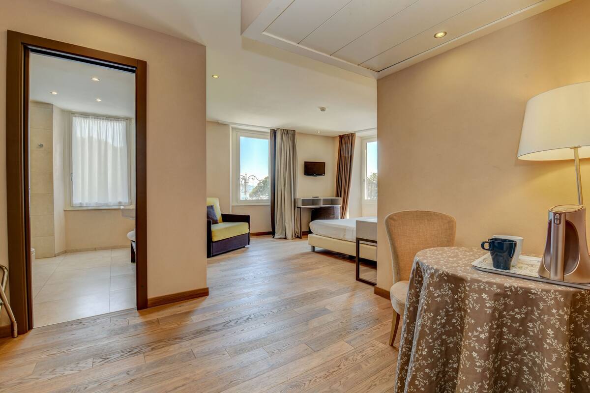 A hotel room with a wooden floor, a bed, a sofa, a table with a lamp, and a TV on the wall.