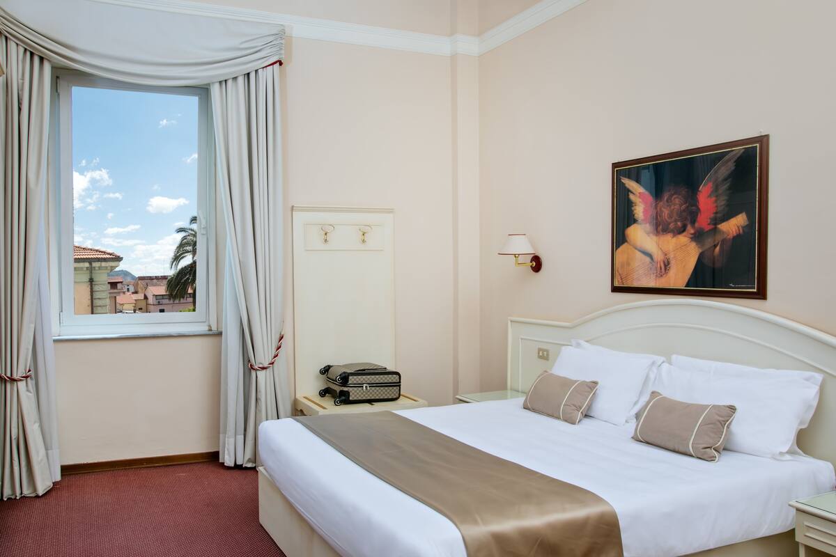 A hotel room with a bed, a nightstand, a lamp, a painting, and a window with curtains.