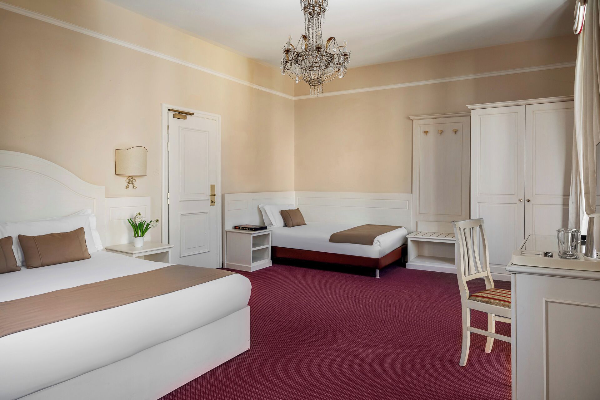 A hotel room with a large bed, a smaller bed, a desk, a chair, a wardrobe, and a chandelier.