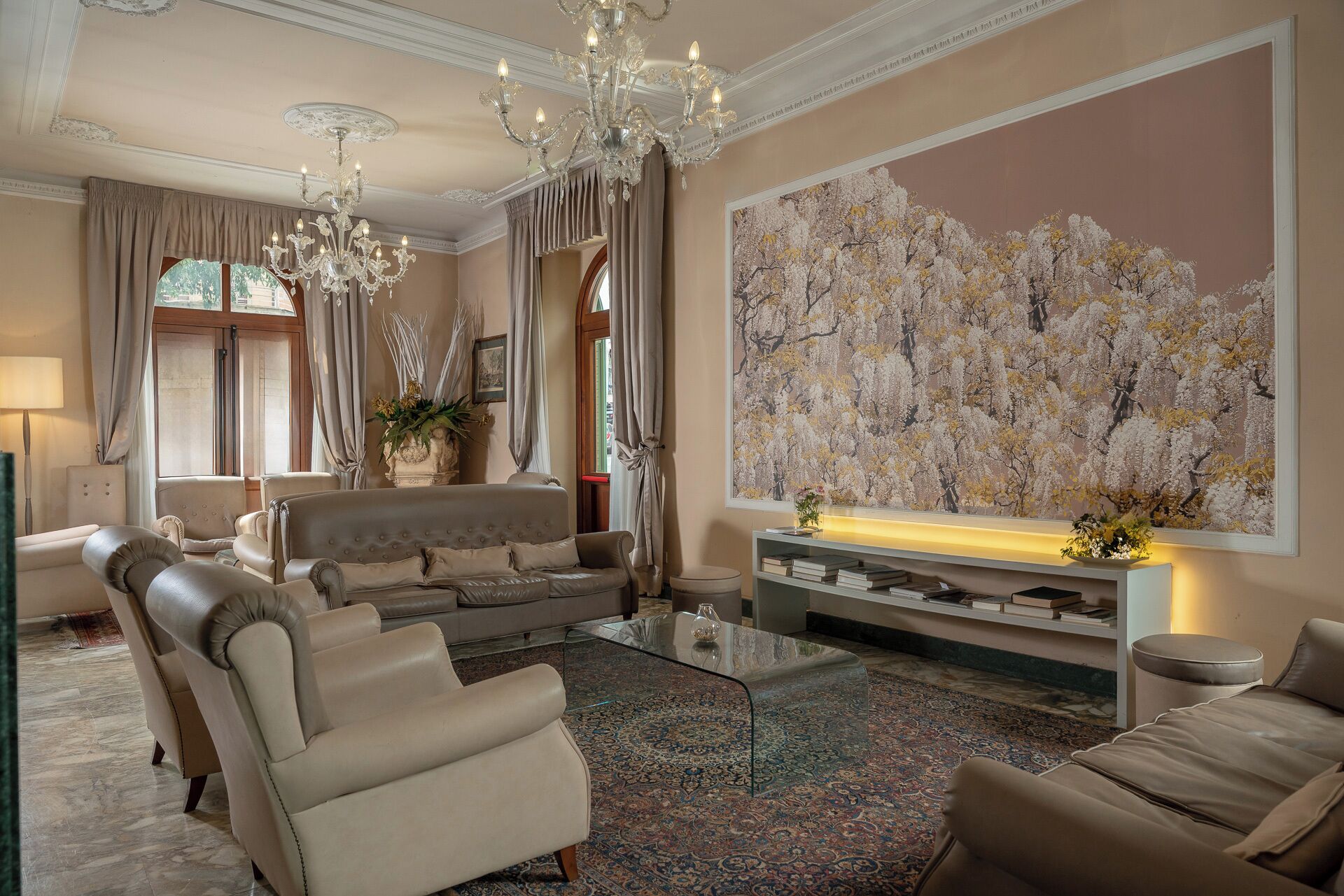 A spacious living room with a large sofa, a glass coffee table, and a chandelier. There is a large floral painting on the wall and a bookshelf with books.