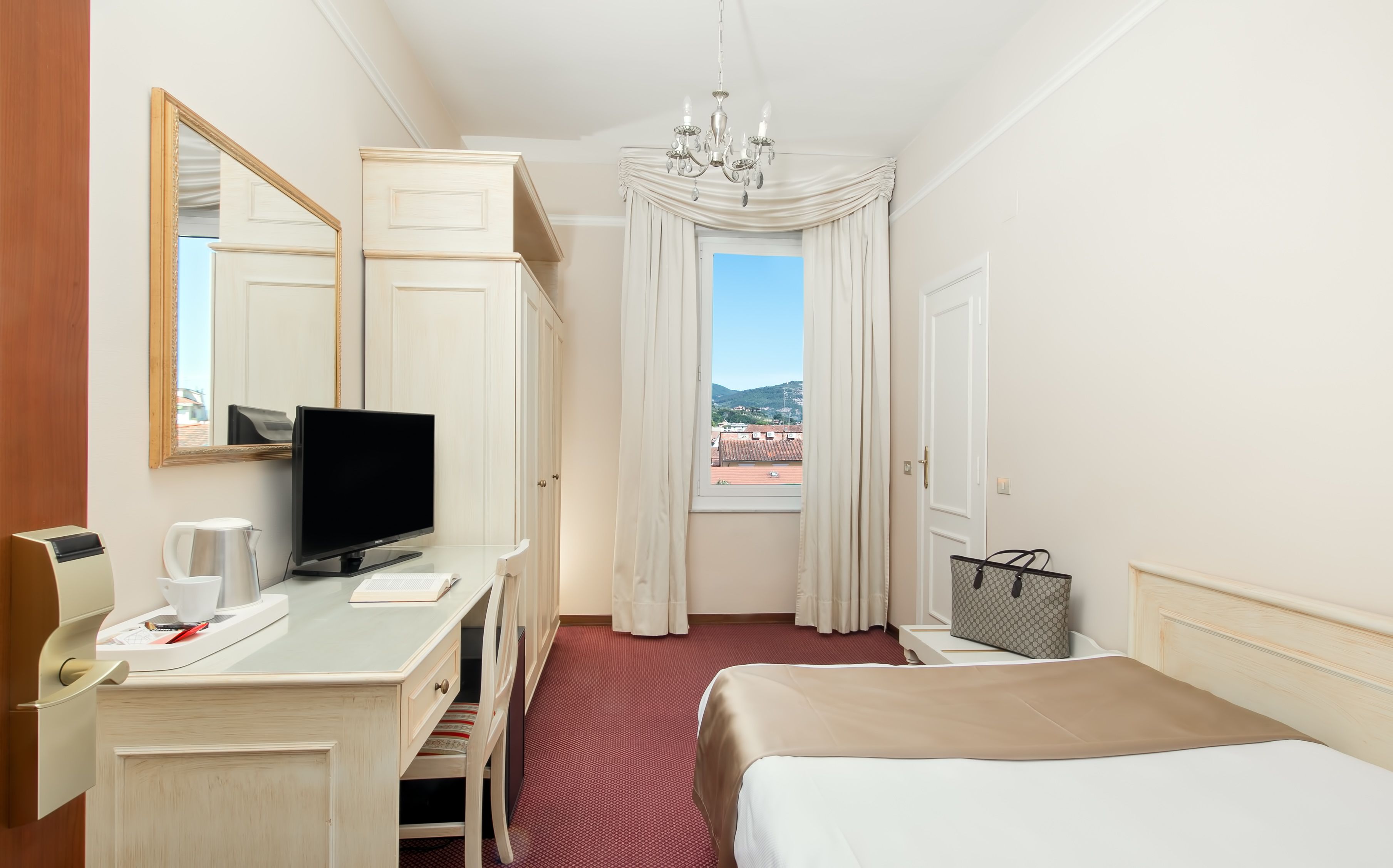 A hotel room with a bed, a desk with a TV, a mirror, a window with curtains, and a view of the outside.