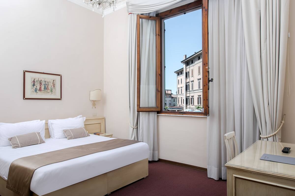 A hotel room with a bed, a desk, a chair, a window with curtains, a picture on the wall, and a view of buildings outside.