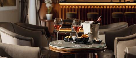 A bar area with two glasses of rosé wine on a round table, a bottle of champagne, olives, and snacks.