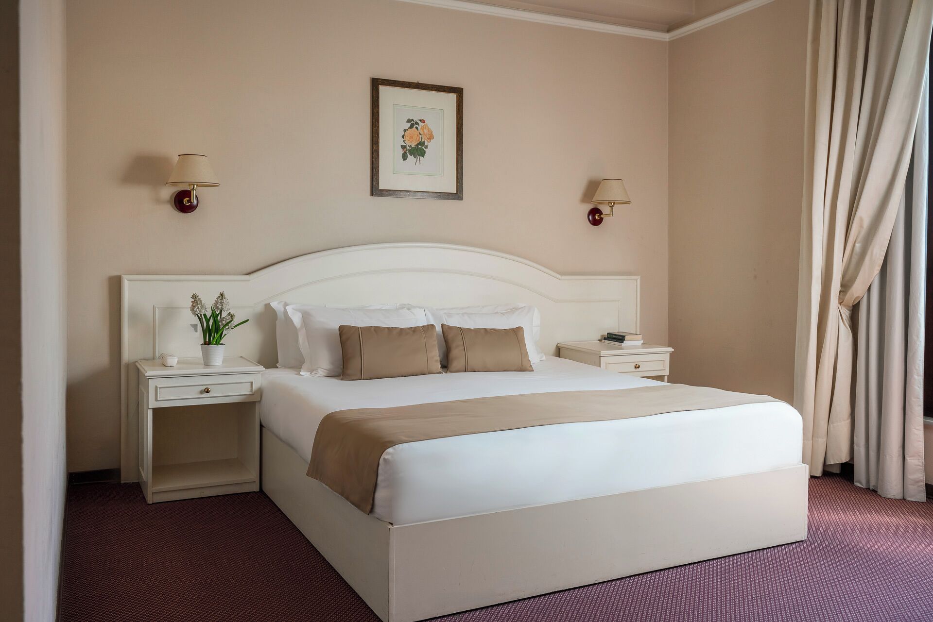 Een hotelkamer met een groot bed, twee nachtkastjes, een lampje en een foto in een lijst aan de muur, plus een raam met gordijnen.