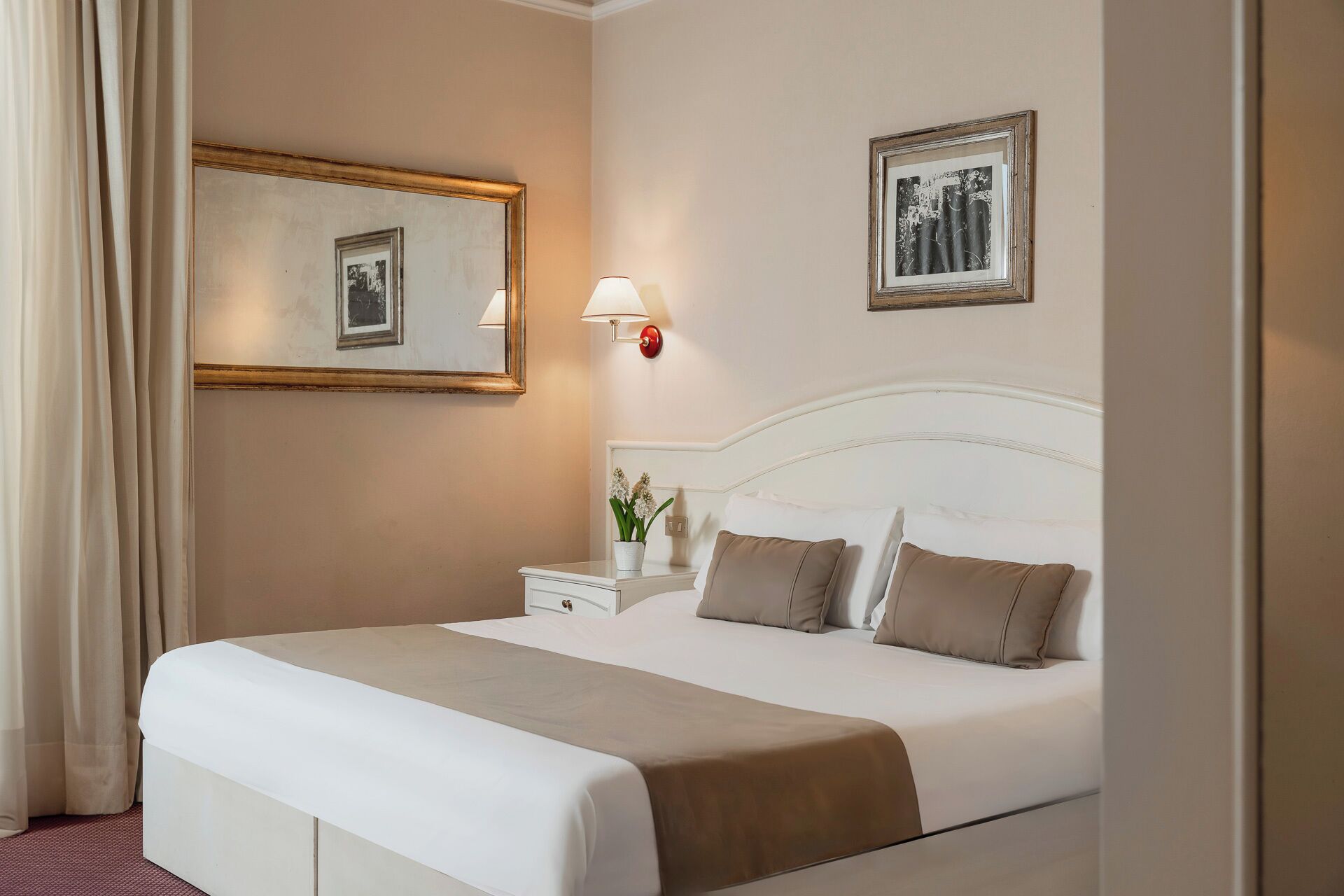 A hotel room with a large bed, a nightstand with a lamp, a mirror, and two framed pictures on the wall.