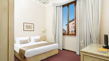 Habitación de hotel con cama, escritorio, silla, ventana con cortinas, candelabro y un cuadro en la pared.