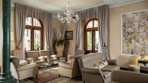 A spacious living room with leather sofas, a glass coffee table, and a chandelier. Large windows with curtains provide natural light, and a large floral painting adorns the wall. - Ercolini E Savi (Montecatini Terme)