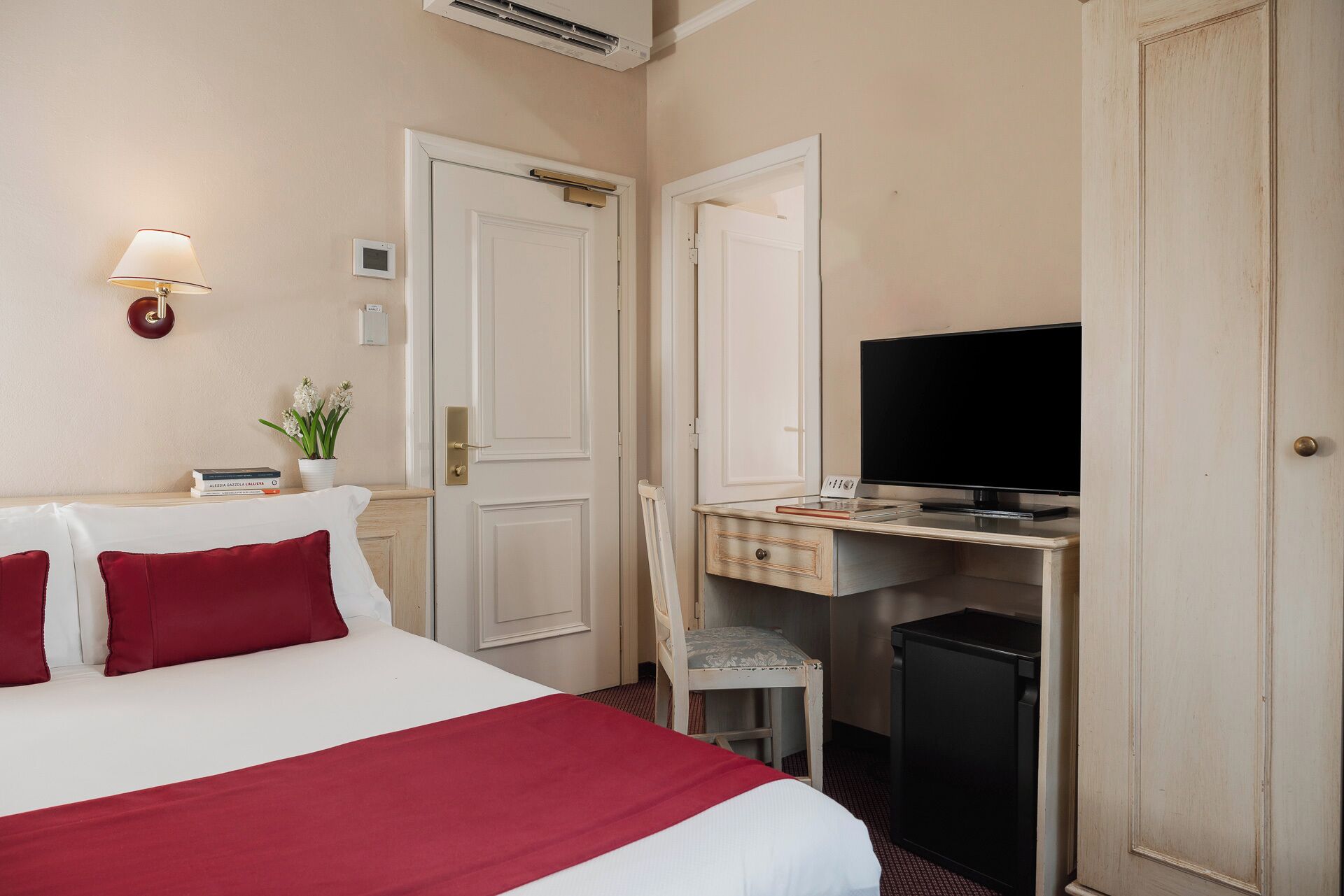 Habitación de hotel con una cama, un escritorio con televisor, una silla y una mesita con planta y libros.
