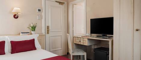 Habitación de hotel con una cama, un escritorio con televisor, una silla y una mesita con planta y libros.