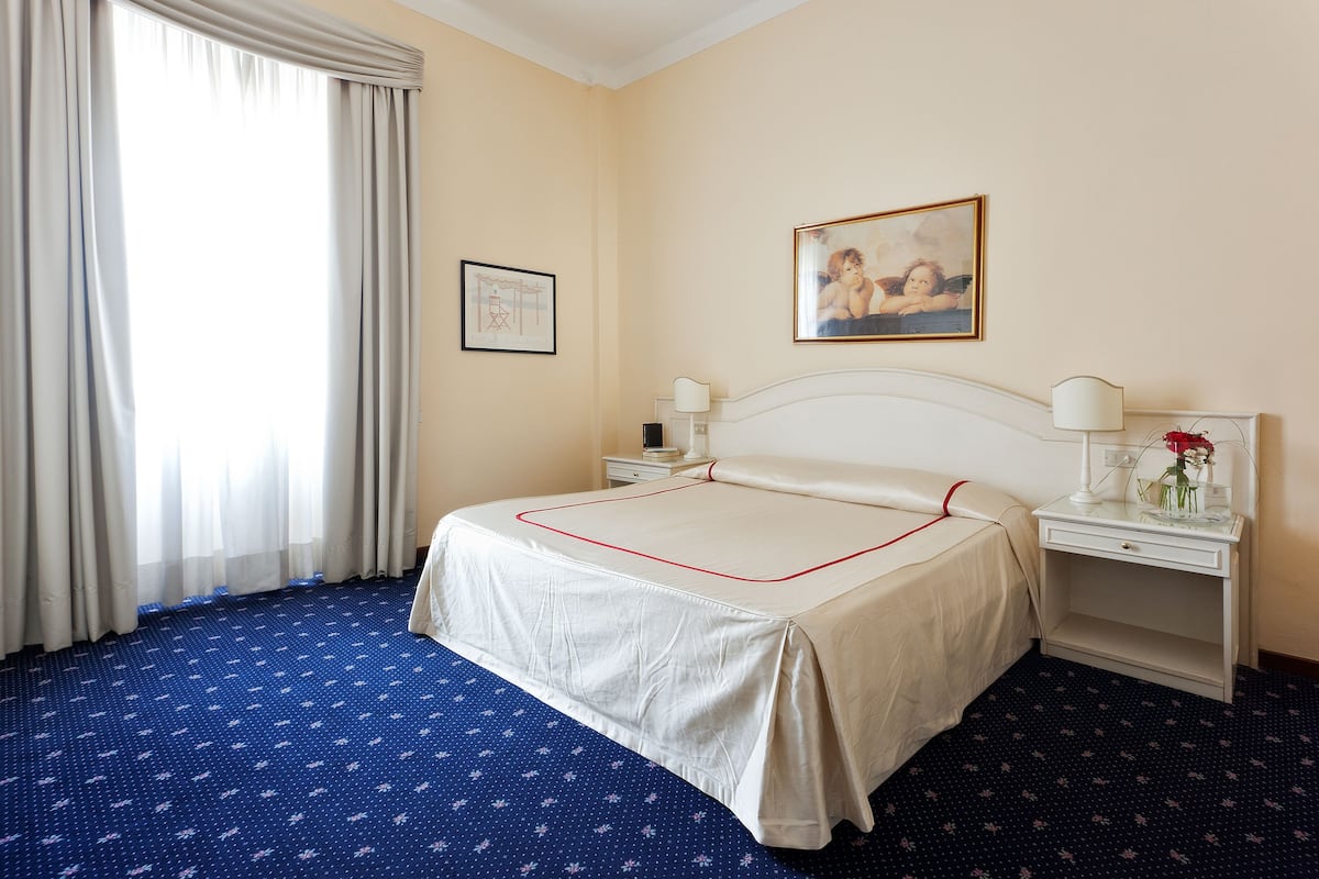 A hotel room with a bed, a nightstand, a lamp, a framed picture on the wall, and a window with curtains.