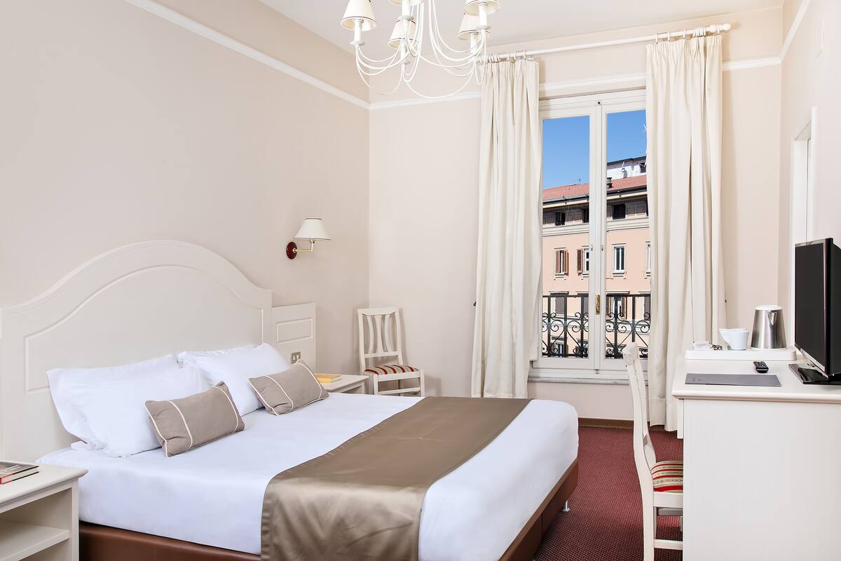 A hotel room with a bed, a desk, a chair, a TV, and a window with a view of buildings.