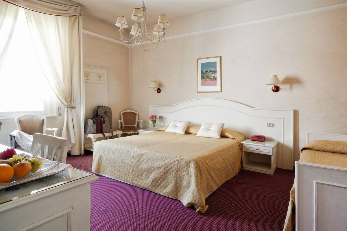 A hotel room with a large bed, a desk with a chair, a small table with fruit, and a window with curtains.