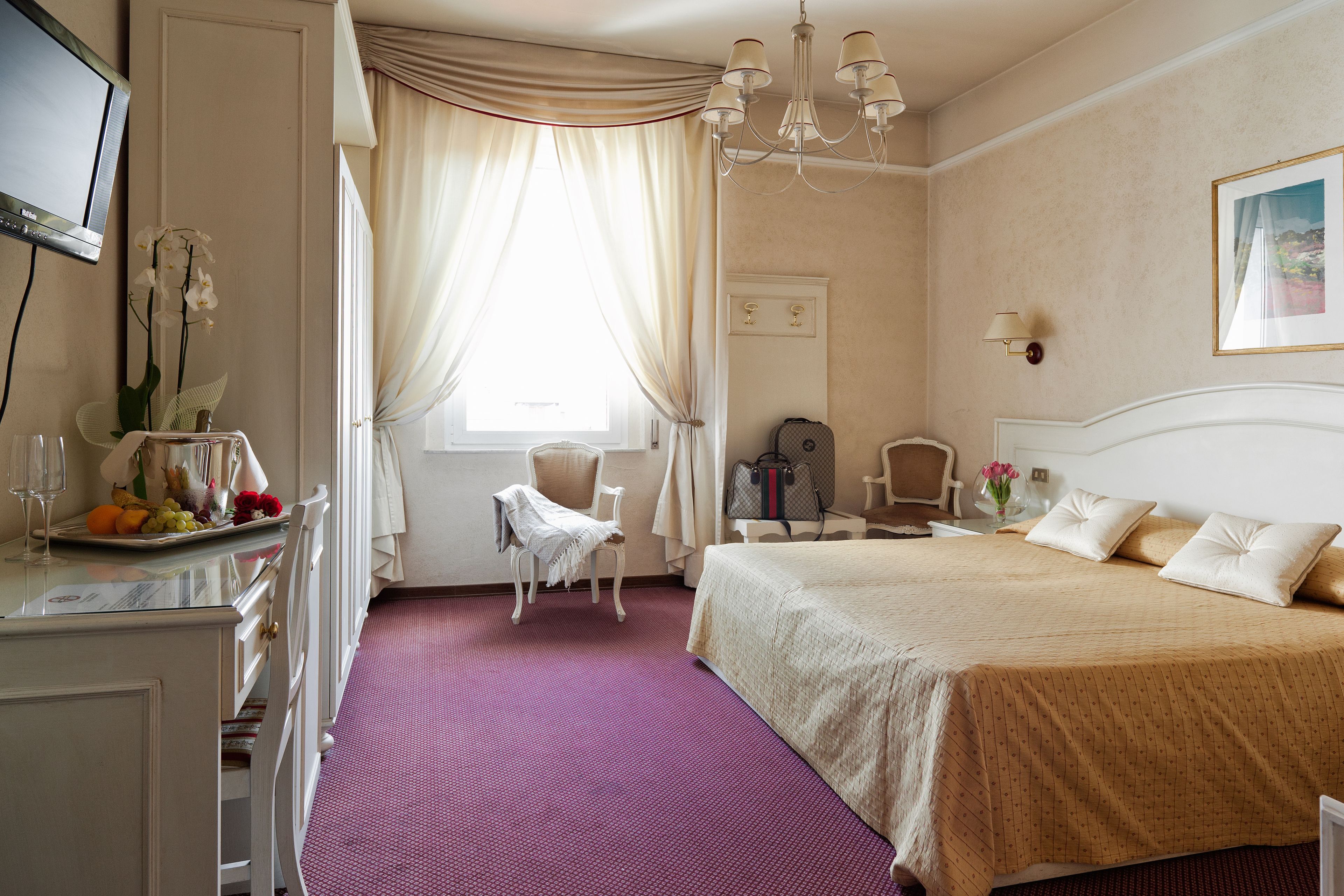 A hotel room with a bed, a desk with a fruit tray, a chair, a TV, and a window with curtains.