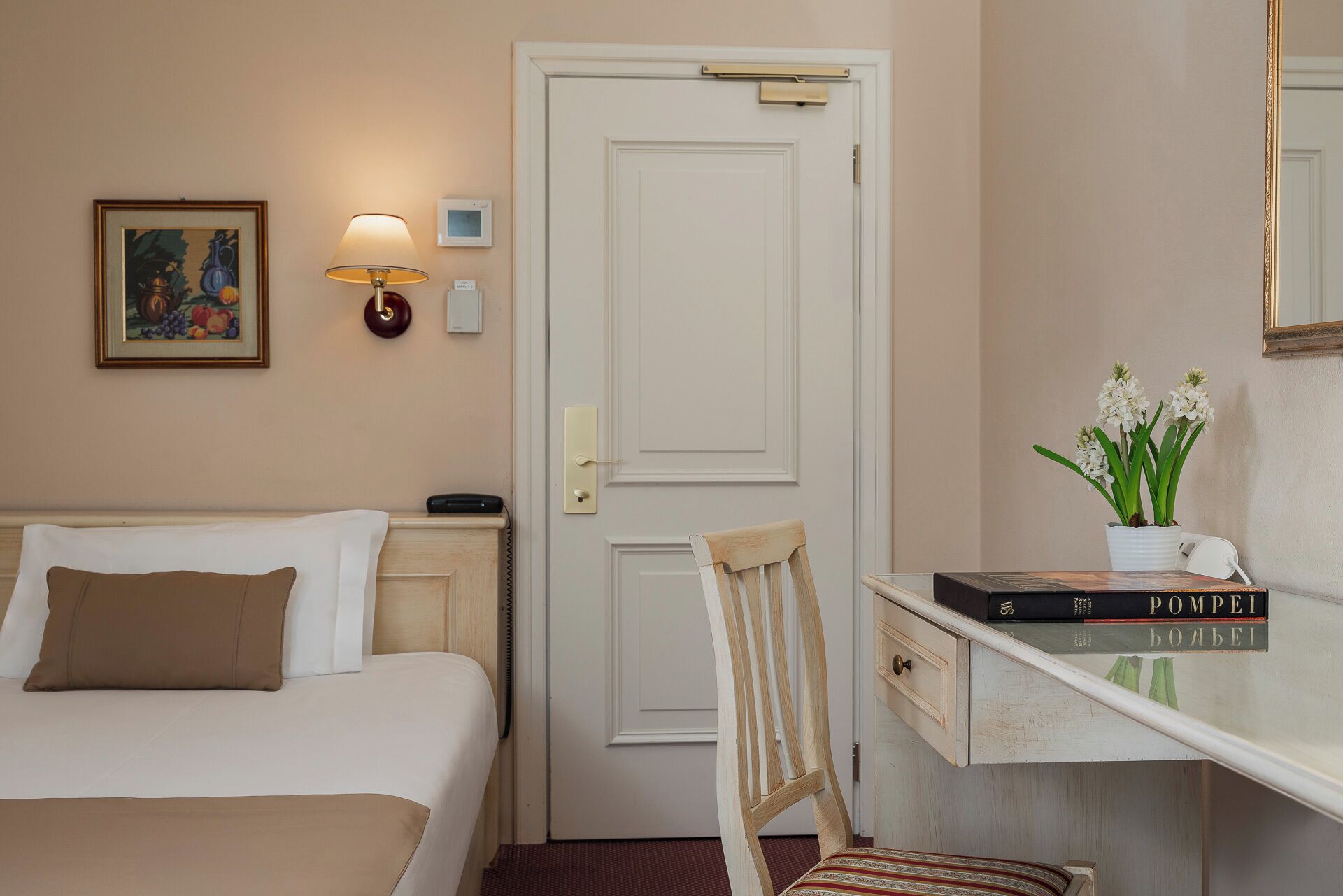 Habitación de hotel con cama, mesita de noche, lámpara, cuadro enmarcado, escritorio con silla, espejo y un jarrón con flores.