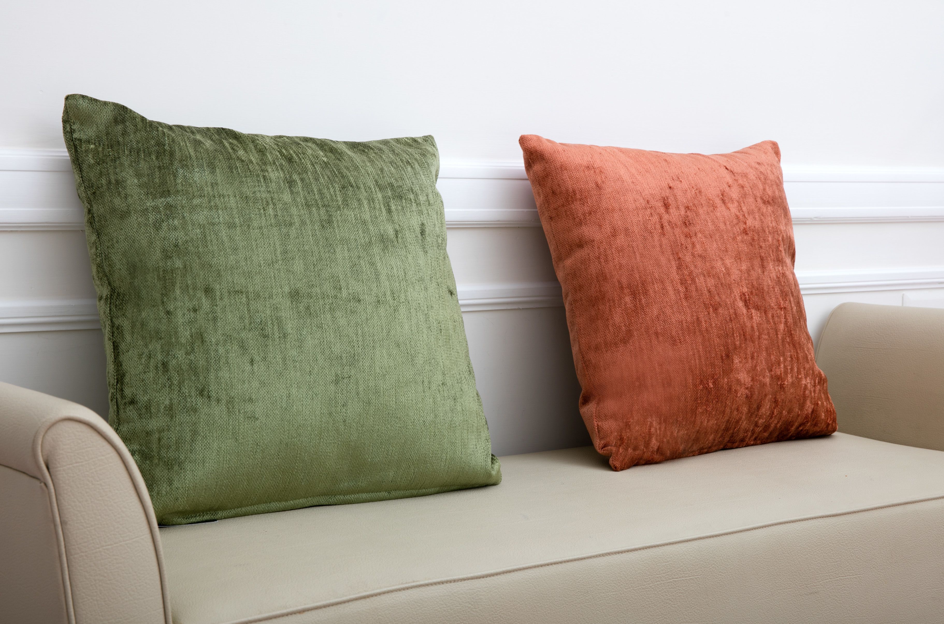 Dos almohadas cuadradas, una verde y otra naranja, sobre un sofá beige frente a una pared blanca.