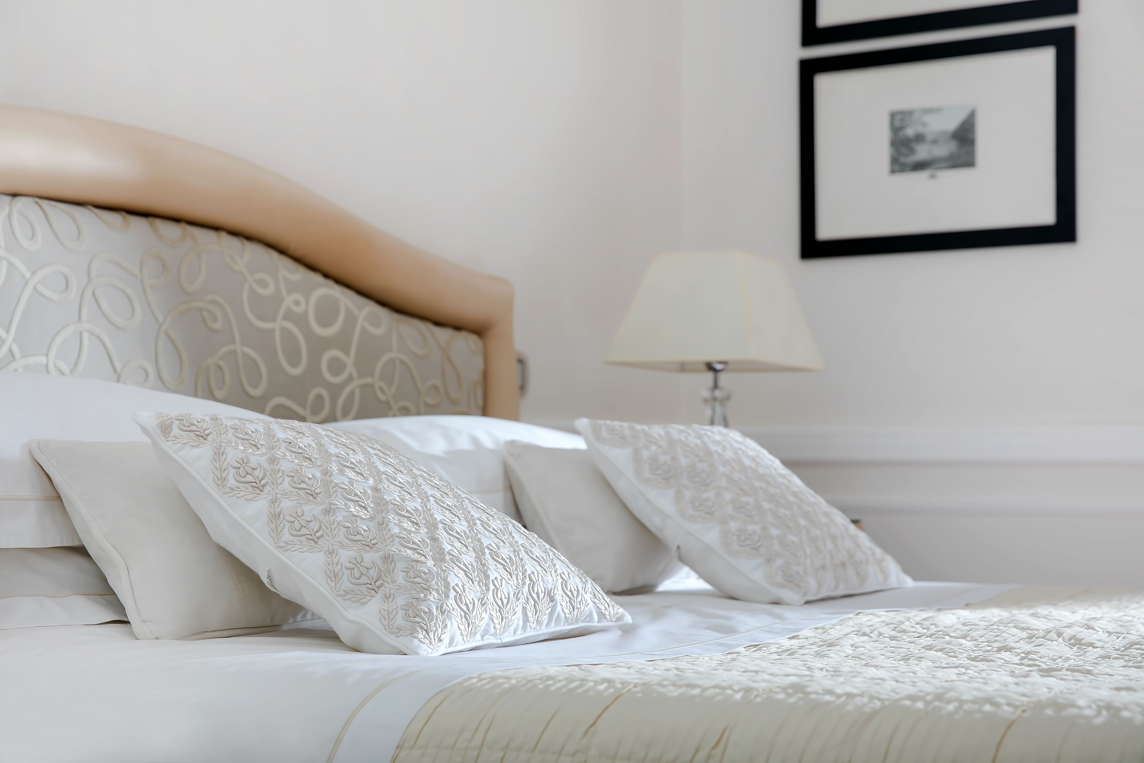Un lit bien fait, avec du linge de lit blanc et des coussins décoratifs, une lampe de chevet et des tableaux encadrés au mur.