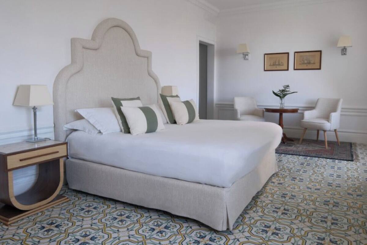 A hotel room with a large bed, a bedside table, a chair, and a small table with a lamp.