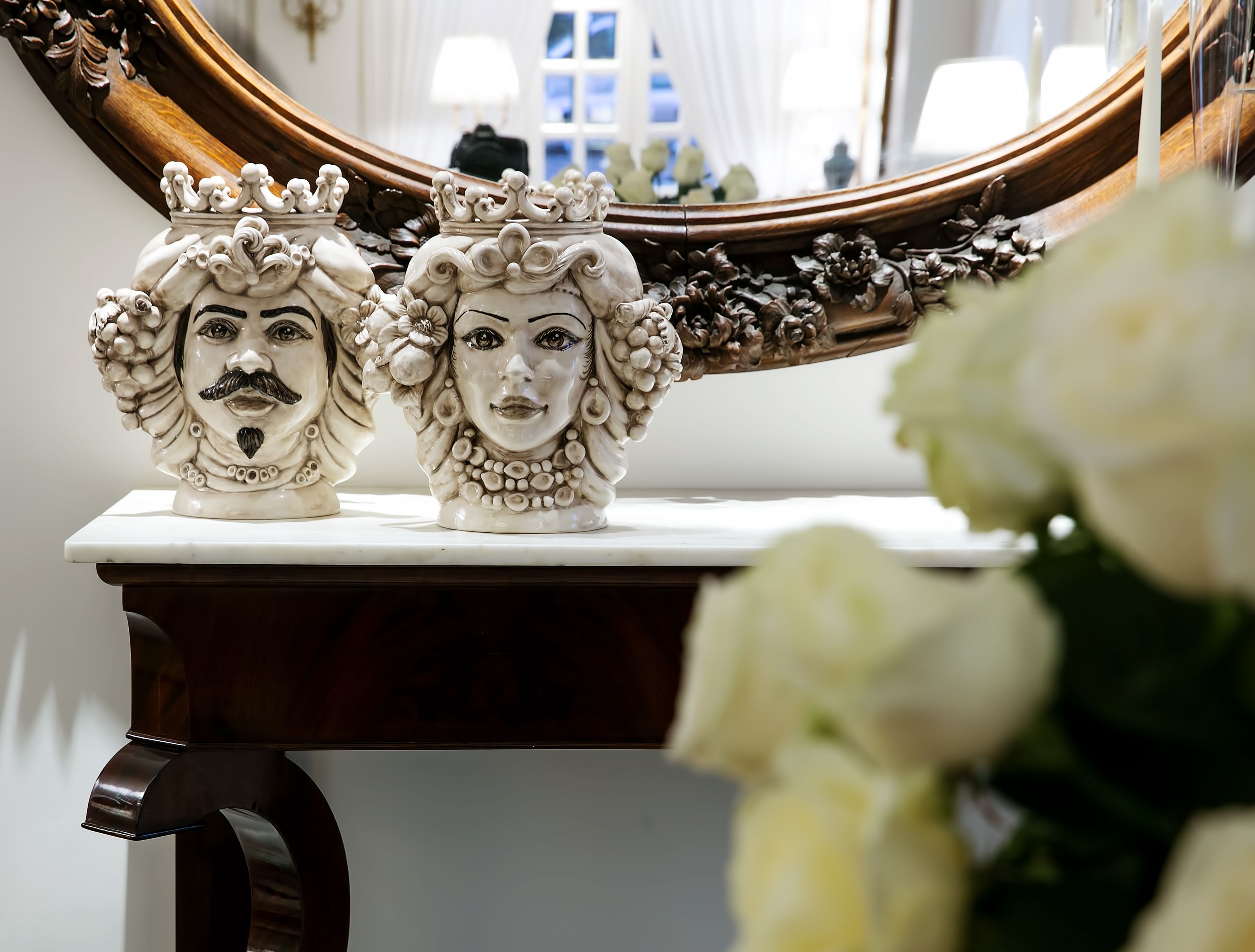 Two ornate ceramic busts on a white marble table with a mirror reflecting a room.