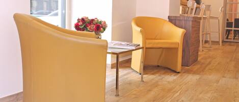 A yellow armchair and a smaller chair in a room with wooden flooring, a table with a vase of flowers, and a desk with chairs in the background.