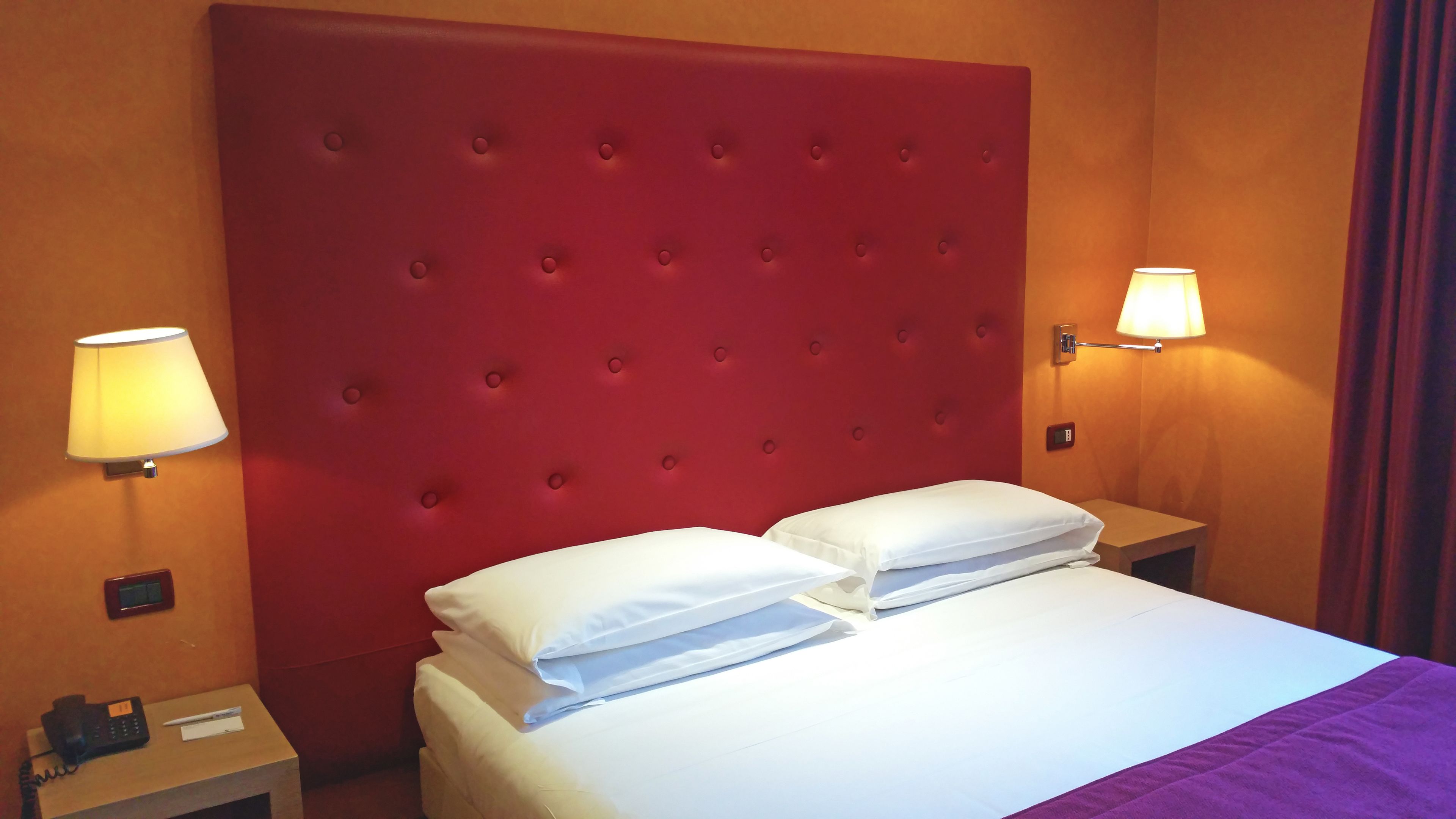 A hotel room with a red tufted headboard, two white pillows, a bed with a purple blanket, a bedside lamp, a phone on the nightstand, and a wall clock.